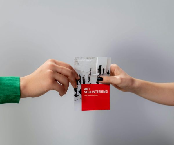 Two hands exchange an art volunteering flyer, emphasizing community and creativity.
