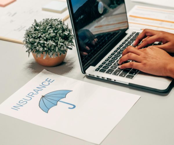 Hands typing on a laptop at a desk with an insurance paper and plant, suggesting a work environment.