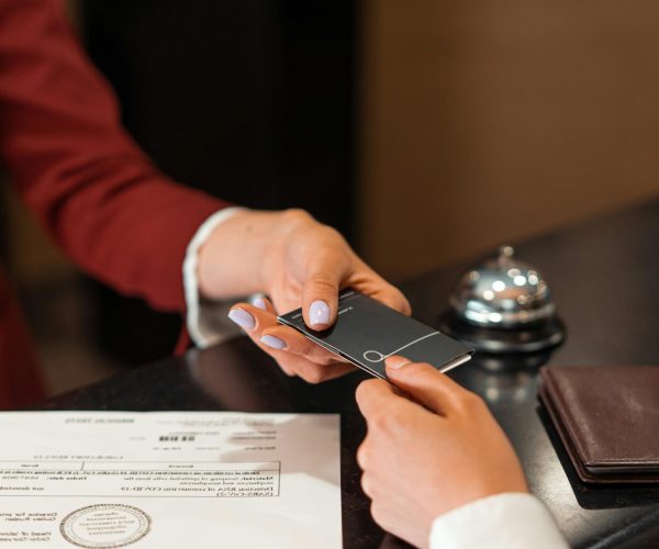 A hotel receptionist hands a key card to a guest over the counter, with documents visible.